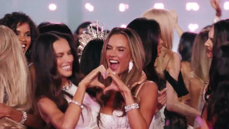 Modelos sorridentes se abraçam no encerramento do desfile; uma delas faz um coração com as mãos, e outra usa uma coroa de cristais