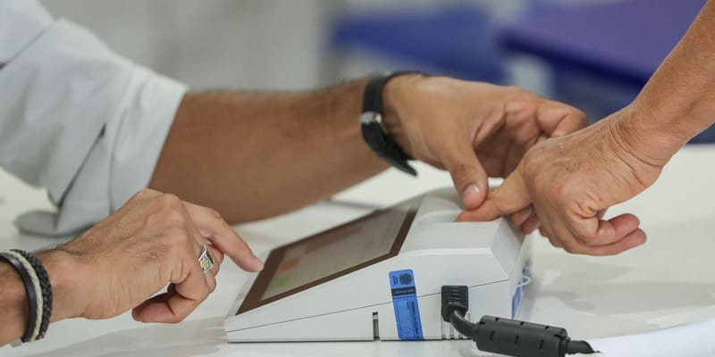 Duas mãos sobre a urna eletrônica, uma delas encostando o dedo no leitor biométrico