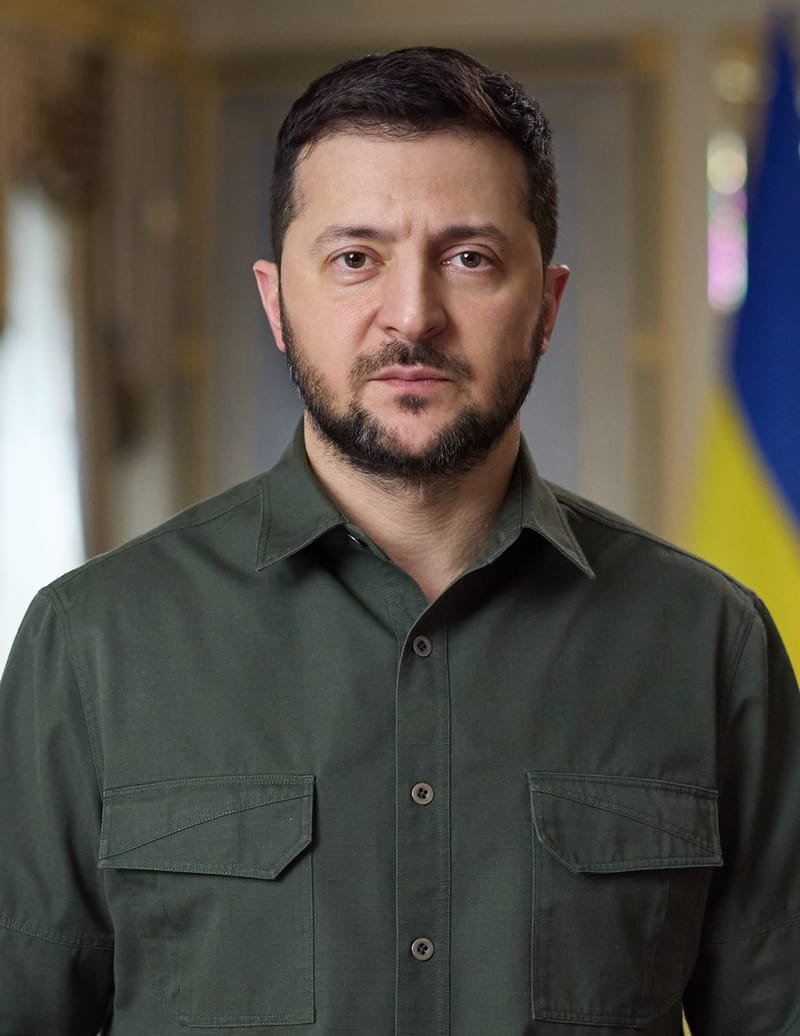 Volodymyr Zelensky, de camisa verde-oliva e sério, com a bandeira da Ucrânia ao fundo, em retrato de frente