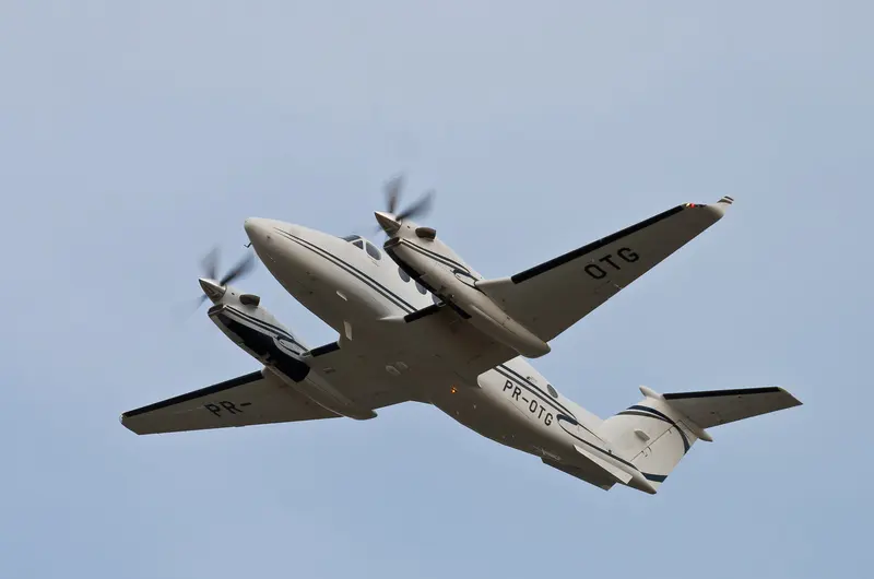 Bimotor executivo turboélice Beechcraft King Air, branco com faixas azuis, logo após decolar, visto de baixo contra o céu