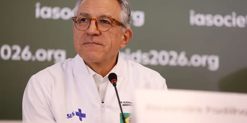 Alexandre Padilha, de óculos de armação marrom e jaqueta branca do SUS, fala diante de um microfone, com painéis da conferência ao fundo