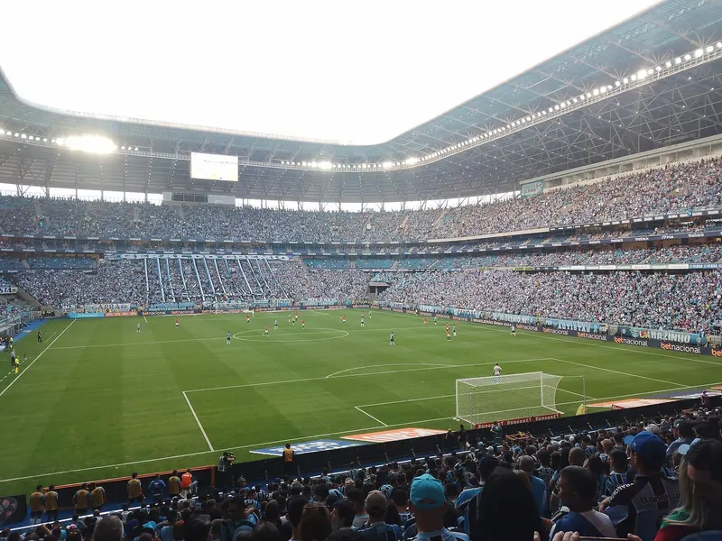 Arquibancadas cheias da Arena do Grêmio, com torcedores de azul em primeiro plano e jogadores em campo, em dia de jogo