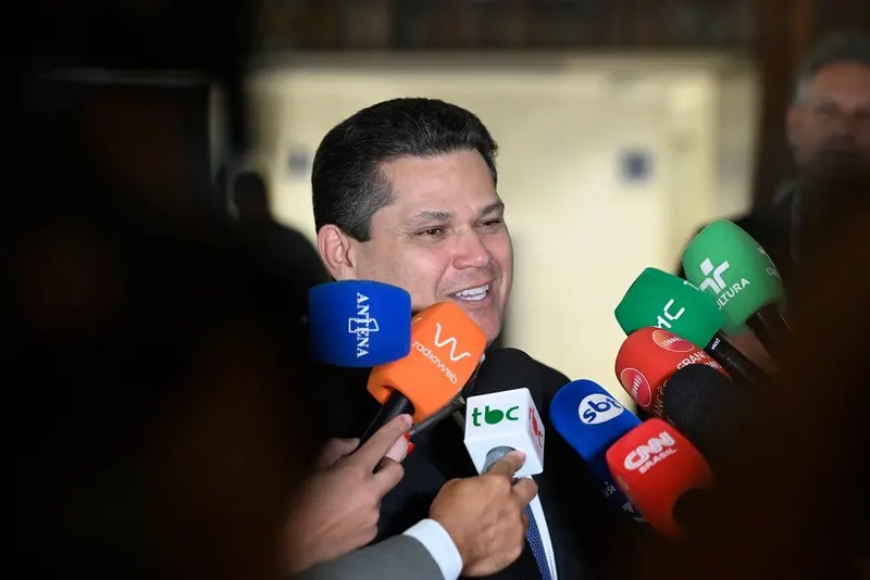 Davi Alcolumbre sorri de terno escuro diante de vários microfones de emissoras, em um ambiente com iluminação escura