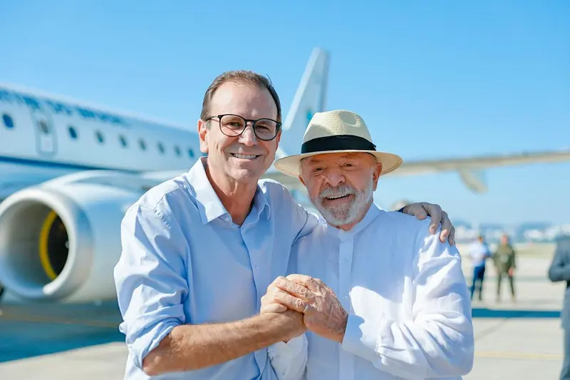 Eduardo Paes, de óculos e camisa azul-clara, abraça Lula, de chapéu de palha e camisa branca, diante de um avião da FAB