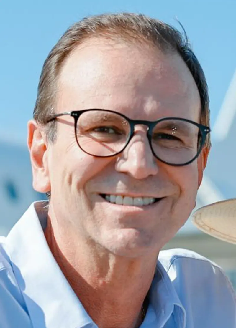 Close do rosto sorridente de Eduardo Paes, de óculos de armação escura e camisa azul-clara, ao ar livre
