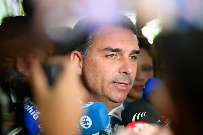 Flávio Bolsonaro, de terno, fala a jornalistas cercado de microfones