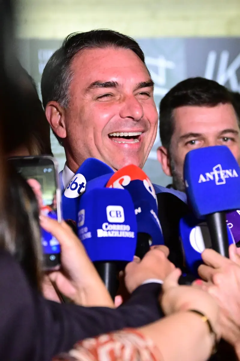 Flávio Bolsonaro sorri de boca aberta cercado de microfones de emissoras de TV