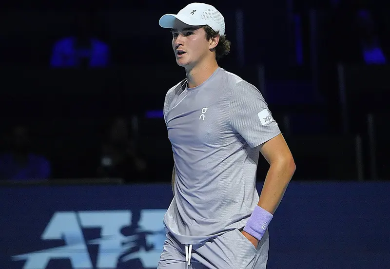 João Fonseca, de boné branco e camiseta cinza, com a mão na cintura, na quadra da Basileia, com o logotipo da ATP ao fundo