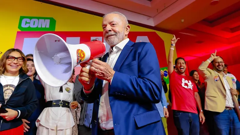 Lula, de blazer azul, segura um megafone e sorri diante de um painel amarelo e vermelho com o nome Lula, cercado de apoiadores