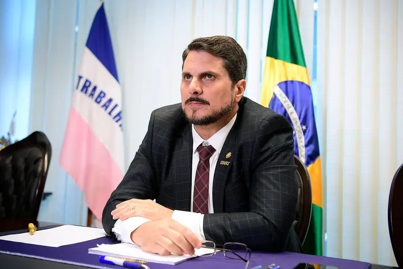 Marcos do Val, de barba, paletó xadrez e gravata vinho, sentado à mesa, com as bandeiras do Brasil e do Espírito Santo ao fundo