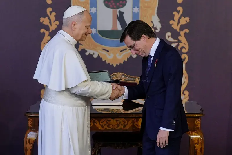 O papa Leão XIV, de batina branca e solidéu, aperta a mão do prefeito de Madri, de terno escuro, diante do brasão da cidade
