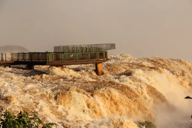 Passarela de metal com grades verdes avança sobre a água barrenta e espumante das Cataratas do Iguaçu em cheia