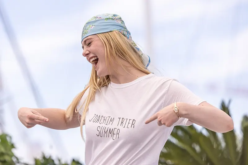 Elle Fanning, de lenço azul estampado na cabeça e camiseta branca, ri e aponta para o próprio peito durante evento em Cannes