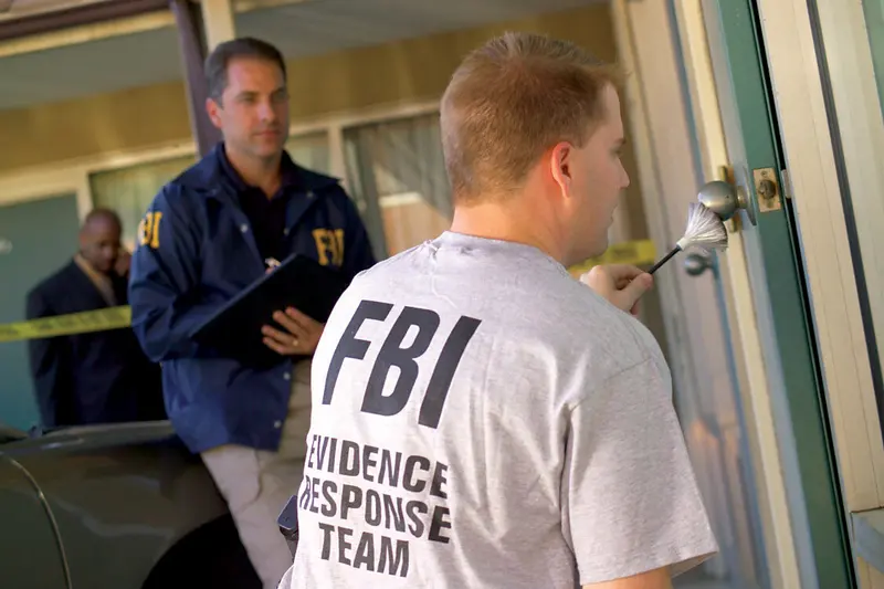 Agentes do FBI, um deles com camiseta da Equipe de Resposta a Evidências, fazem perícia na porta de uma residência isolada por fita