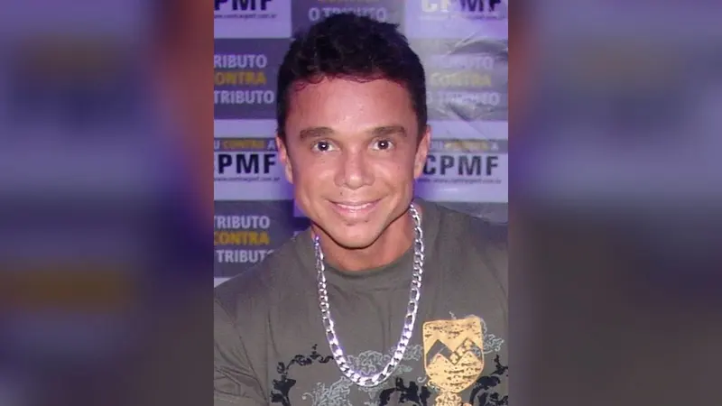 O cantor Netinho sorri, com corrente prateada no pescoço, diante de painel de evento, em 2007