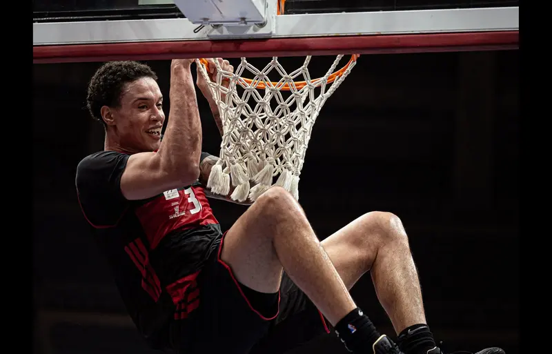 Jogador do Flamengo pendurado no aro depois de uma enterrada, com a camisa rubro-negra, em quadra do time de basquete