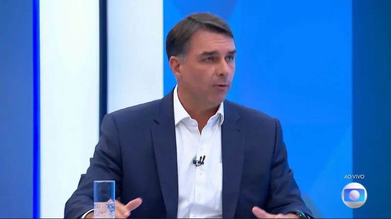 Flávio Bolsonaro, de terno azul-marinho e camisa branca, gesticula durante entrevista ao vivo no estúdio da TV Globo