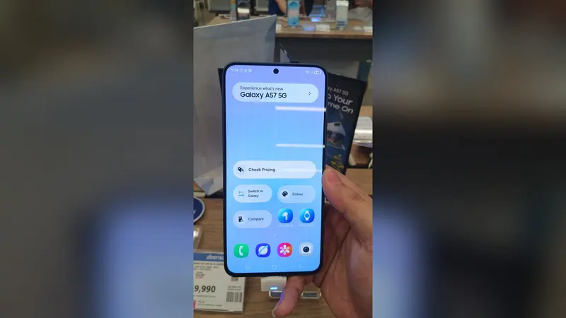 Celular Samsung Galaxy A57 5G em exposição numa loja, seguro por uma mão, com a tela inicial acesa