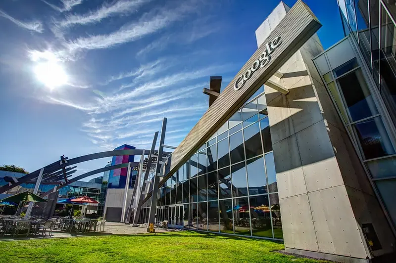 Prédio de vidro e concreto da sede do Google, com o nome da empresa na viga inclinada, sob céu azul