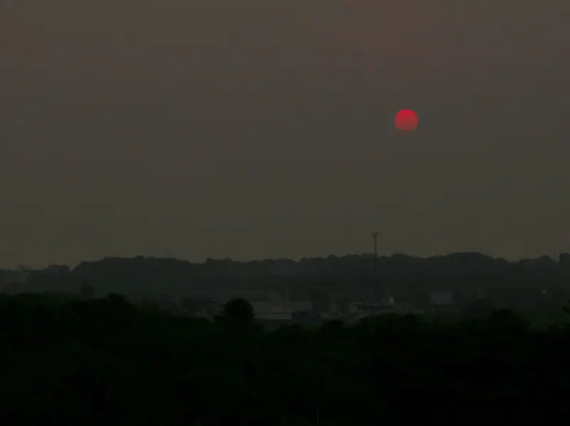 Sol vermelho aparece através de uma camada densa de fumaça sobre Manaus, com prédios e mata escurecidos abaixo