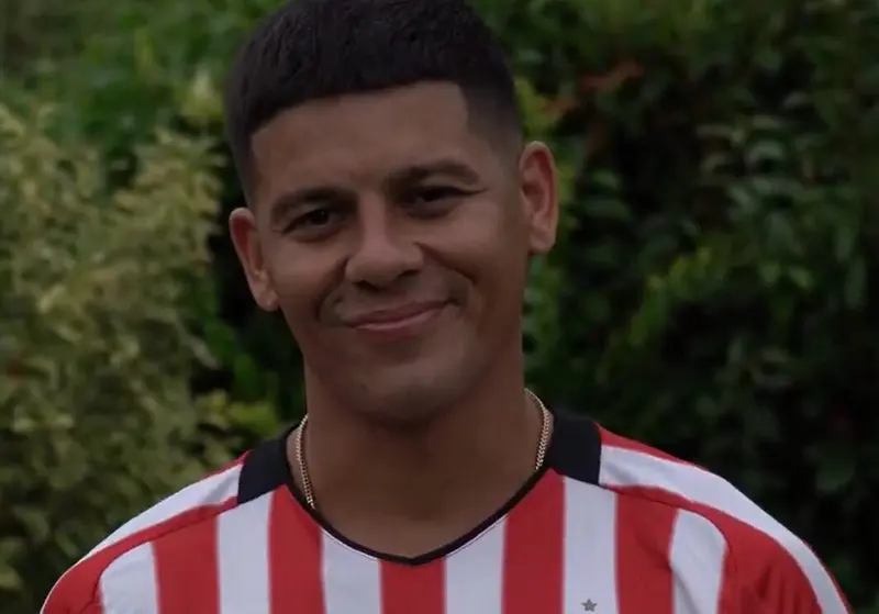 Marcos Rojo sorri vestindo a camisa listrada em vermelho e branco do Estudiantes, com plantas ao fundo