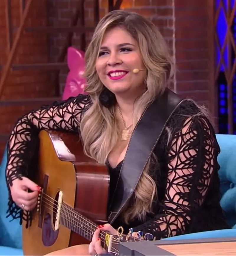 Marília Mendonça sorri sentada em um sofá azul de estúdio de TV, com um violão no colo
