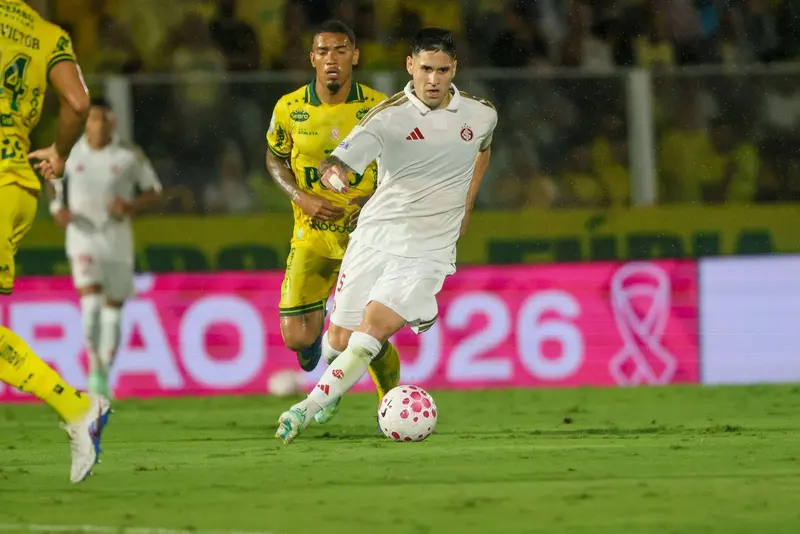 Jogador do Internacional, de camisa branca, conduz a bola pelo gramado enquanto um adversário do Mirassol, de amarelo, acompanha a jogada; ao fundo, placa rosa do Brasileirão