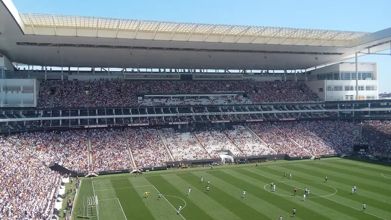 Interior da Neo Química Arena em dia de jogo, com as arquibancadas lotadas, a cobertura branca e o gramado sob céu azul