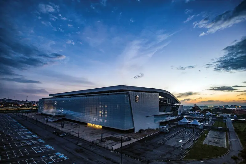 A Neo Química Arena ao entardecer, com a fachada de vidro iluminada, o escudo do Corinthians e o estacionamento vazio à frente, sob céu azul com nuvens