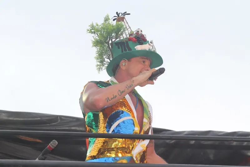 Netinho, de chapéu enfeitado e colete brilhante, canta e estende o braço para o público no alto de um trio elétrico
