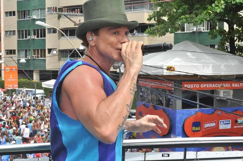 Netinho, de chapéu verde e regata azul, canta ao microfone no alto de um trio elétrico, com o público ao fundo
