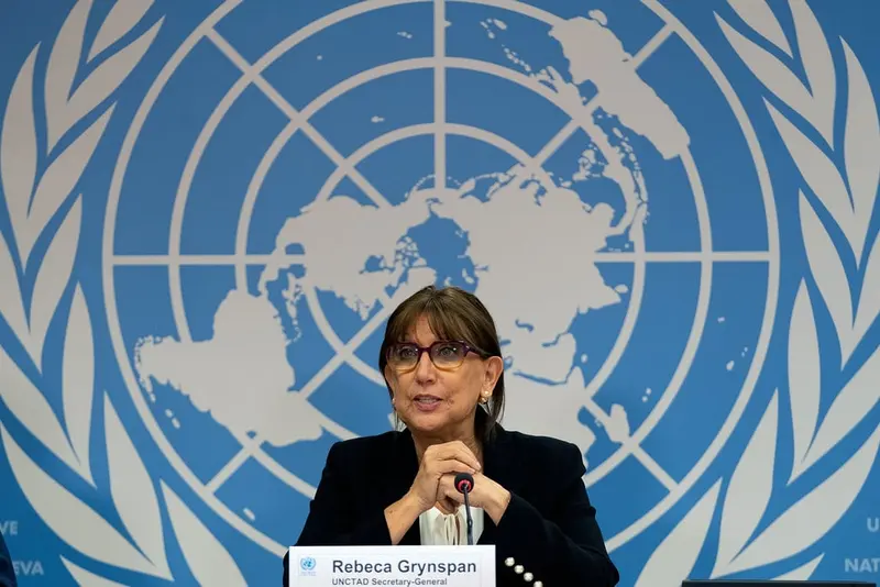 Rebeca Grynspan, de blazer escuro e óculos, fala ao microfone diante do emblema azul e branco da ONU, com a placa de identificação à frente