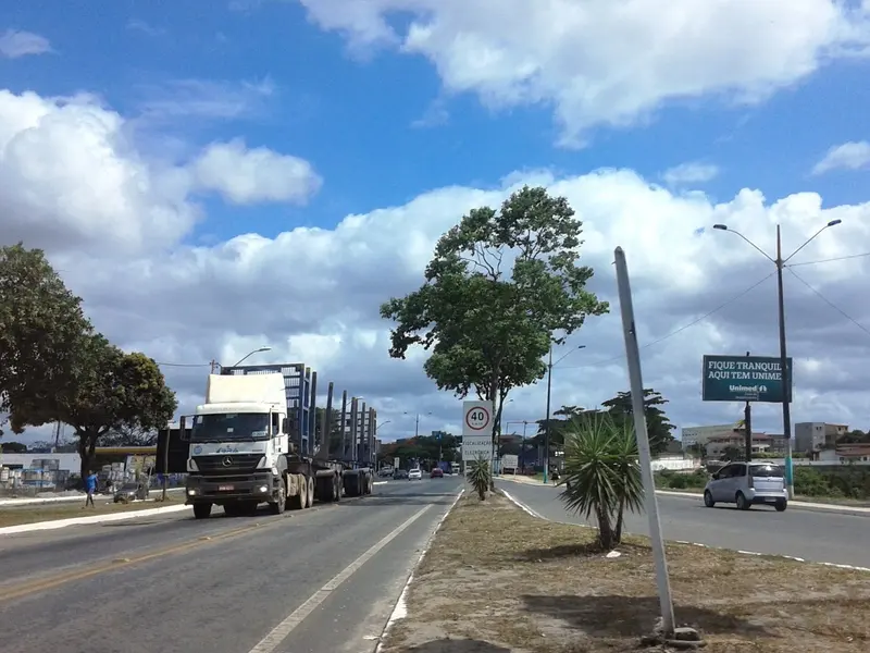 Caminhão de transporte de toras e carros trafegam pelo trecho urbano da BR-101 em Eunápolis, sob céu azul com nuvens