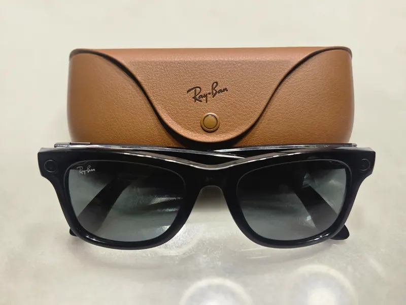Óculos Ray-Ban Meta de armação preta, com câmera discreta no canto da armação, apoiados diante do estojo marrom da marca