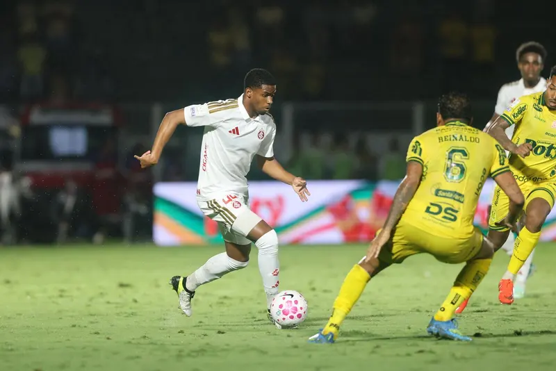 Jogador do Internacional, de uniforme branco, conduz a bola pelo gramado enquanto dois jogadores do Mirassol, de amarelo, tentam a marcação