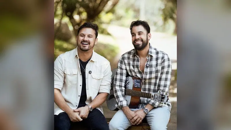 Os sertanejos Clayton e Romário, sorridentes, sentados lado a lado; um deles segura um violão