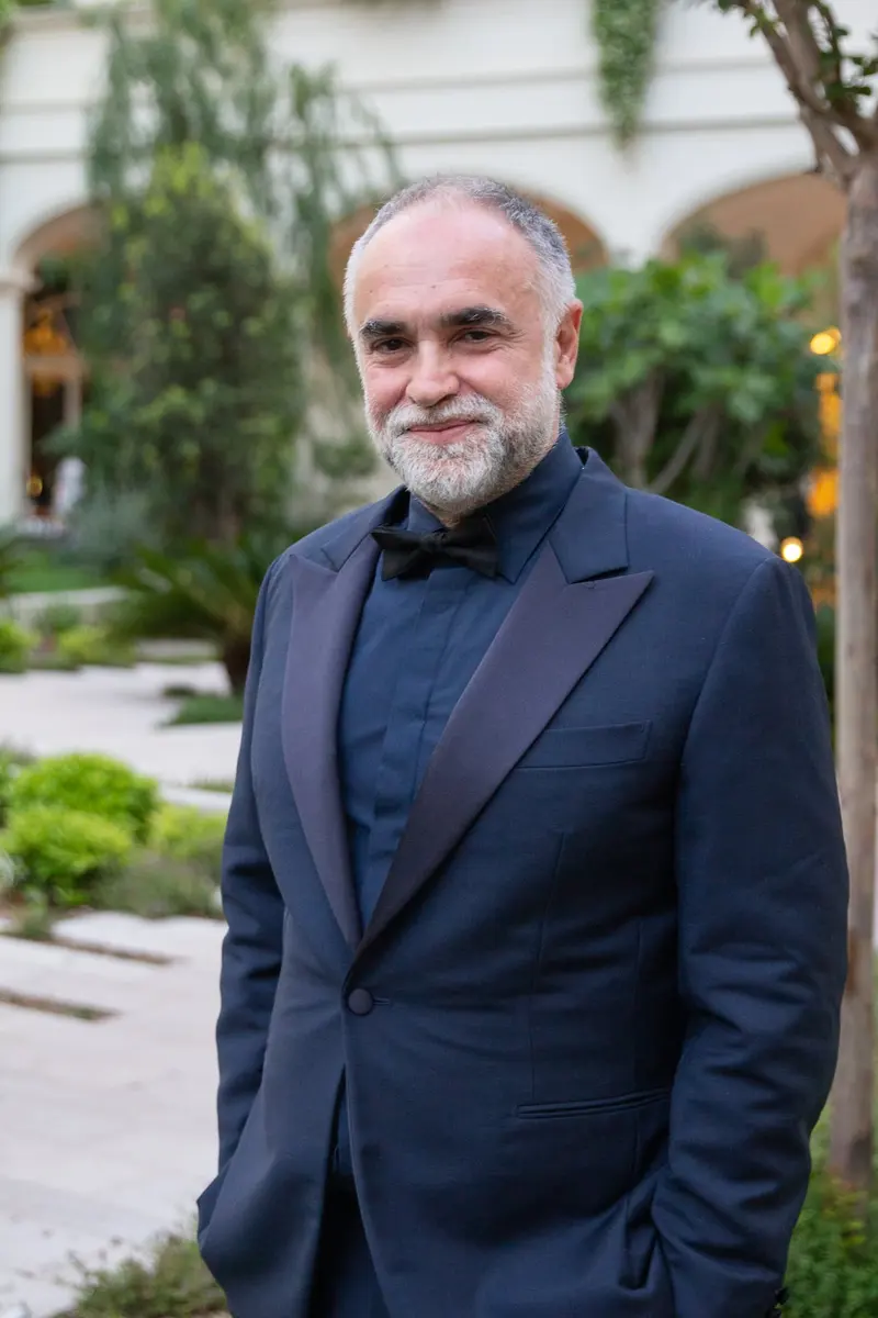 Karim Aïnouz, de barba grisalha, smoking azul-marinho e gravata-borboleta preta, sorri num jardim