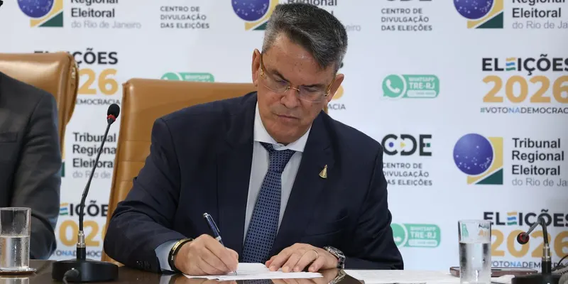 Presidente do TRE-RJ assina documento em mesa, diante de painel do Centro de Divulgação das Eleições 2026