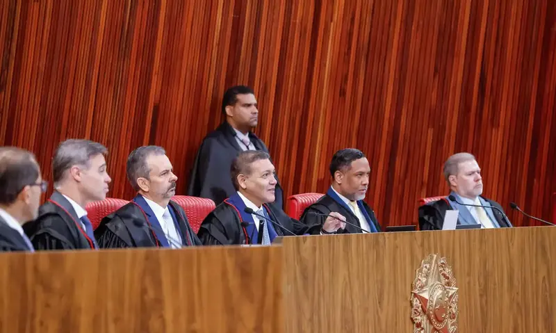 Ministros do TSE, de toga preta, sentados lado a lado na bancada de madeira do plenário, com o painel de madeira ao fundo