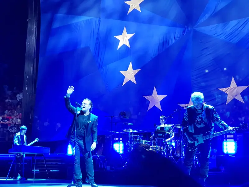 Bono acena ao público ao lado dos integrantes do U2 durante show, sob luz azul
